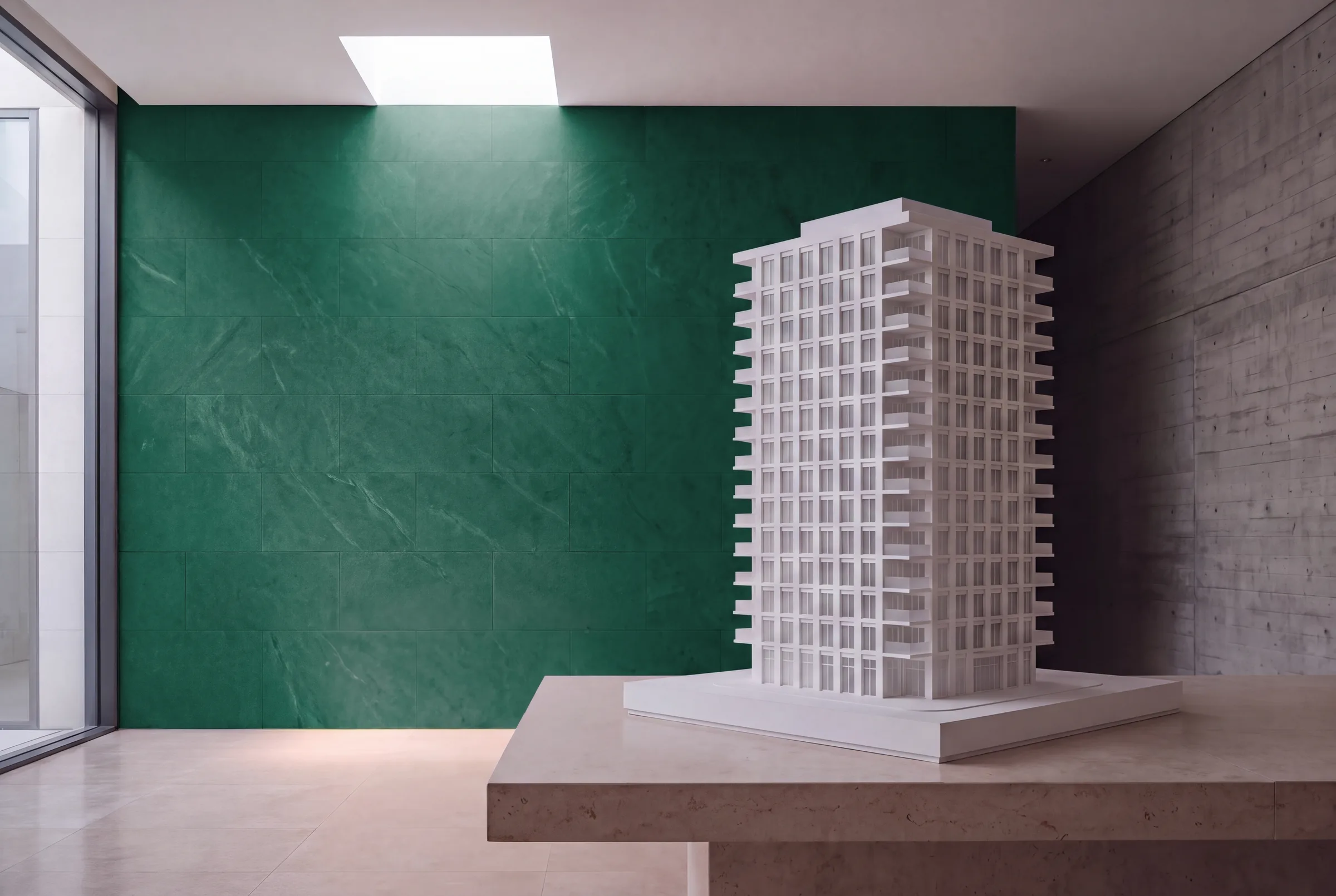 Maqueta blanca de una torre residencial sobre una mesa de piedra, contra un muro de piedra verde en una sala de ventas iluminada por un lucernario